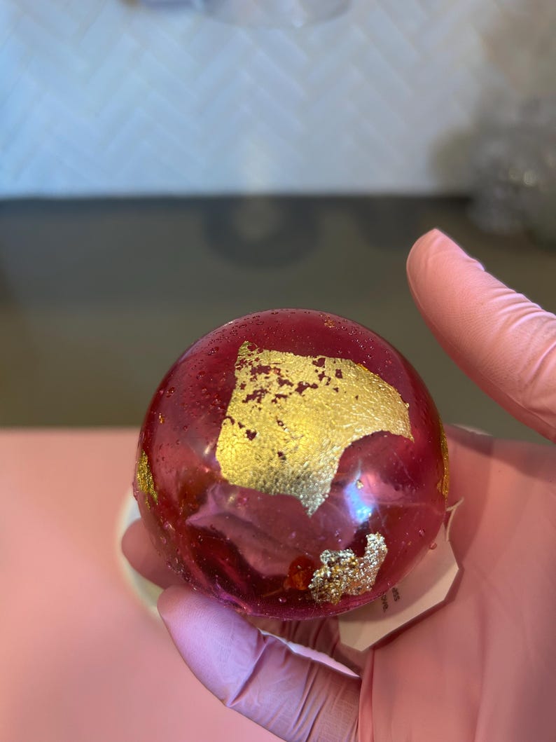 May include: A round, translucent, red object with gold leaf accents. The object is held in a gloved hand. The gold leaf is irregularly shaped and adds a decorative element to the red sphere.