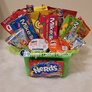 May include: A green basket filled with a variety of snacks, including Mike and Ike candies, Takis chips, Capri Sun juice boxes, and Pringles potato chips. The basket is decorated with green tissue paper and red ribbons. The word "Nerds" is visible on a candy box.