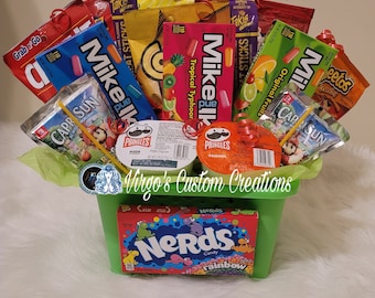 May include: A green basket filled with a variety of snacks, including Mike and Ike candies, Takis chips, Capri Sun juice boxes, and Pringles potato chips. The basket is decorated with green tissue paper and red ribbons. The word "Nerds" is visible on a candy box.