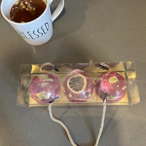 May include: A white mug with the word "BLESSED" in black lettering sits next to a clear package containing three pink bath bombs. The bath bombs have gold accents and dried botanicals. One bath bomb has a dried citrus slice.