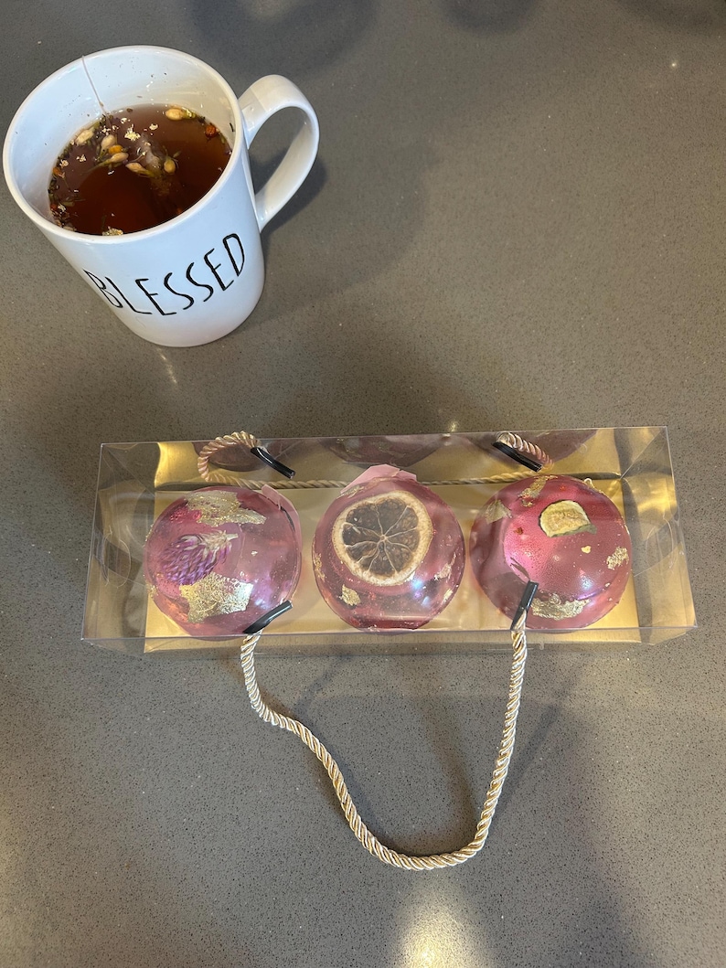 May include: A white mug with the word "BLESSED" in black lettering sits next to a clear package containing three pink bath bombs. The bath bombs have gold accents and dried botanicals. One bath bomb has a dried citrus slice.