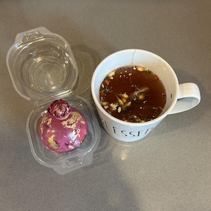 May include: A clear plastic container holds a pink, spherical dessert with gold leaf accents and a flower on top. Next to it is a white mug filled with tea, with the word "BLESSED" visible on the side.