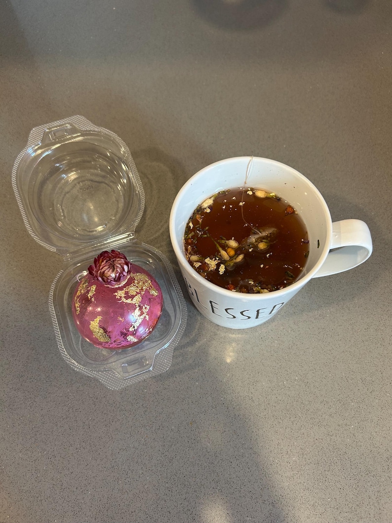 May include: A clear plastic container holds a pink, spherical dessert with gold leaf accents and a flower on top. Next to it is a white mug filled with tea, with the word "BLESSED" visible on the side.