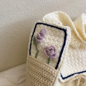 Peut inclure: Un sac cabas en crochet blanc avec une poche beige et un motif floral. Les fleurs sont violettes et vertes et sont crochet&eacute;es sur le sac.