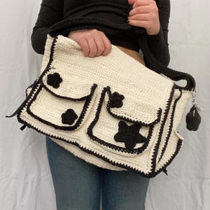 Pu&ograve; includere: Una borsa bianca all'uncinetto con dettagli in crochet nero. La borsa ha tre tasche con decorazioni a forma di fiore e stella nere. La borsa ha una tracolla in crochet nero.