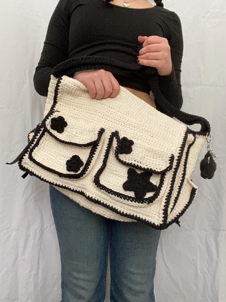 Pu&ograve; includere: Una borsa bianca all'uncinetto con dettagli in crochet nero. La borsa ha tre tasche con decorazioni a forma di fiore e stella nere. La borsa ha una tracolla in crochet nero.