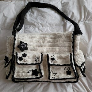 Pu&ograve; includere: Una borsa tote in crochet bianco con bordi neri e stelle ricamate. La borsa ha pi&ugrave; tasche e una tracolla in crochet nero.