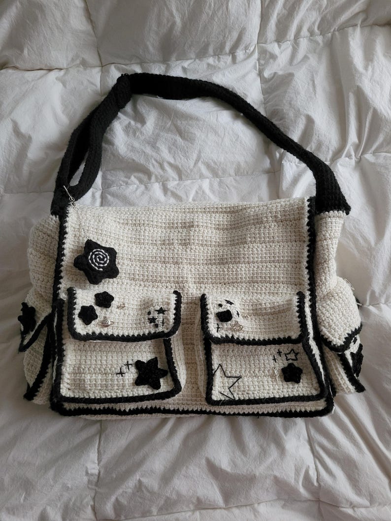Pu&ograve; includere: Una borsa tote in crochet bianco con bordi neri e stelle ricamate. La borsa ha pi&ugrave; tasche e una tracolla in crochet nero.
