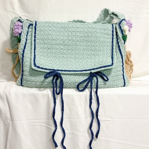 Peut inclure: Un sac en crochet bleu clair avec une bordure bleu marine et deux longues attaches bleu marine. Le sac a une bandouli&egrave;re en crochet marron et est d&eacute;cor&eacute; de petites fleurs en crochet violet et blanc.