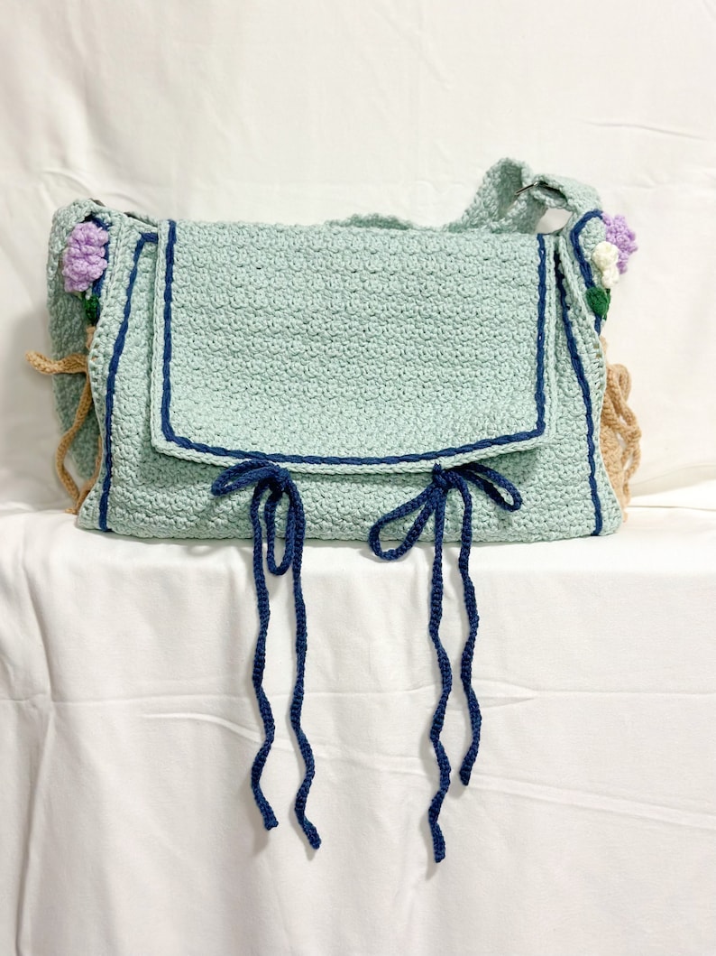 Peut inclure: Un sac en crochet bleu clair avec une bordure bleu marine et deux longues attaches bleu marine. Le sac a une bandouli&egrave;re en crochet marron et est d&eacute;cor&eacute; de petites fleurs en crochet violet et blanc.