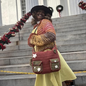 Pu&ograve; includere: Una donna indossa un cardigan di uncinetto multicolore, un vestito giallo e un cappello marrone. Porta una borsa di uncinetto con decorazioni floreali. La borsa ha due tasche grandi e una tasca pi&ugrave; piccola sul davanti.