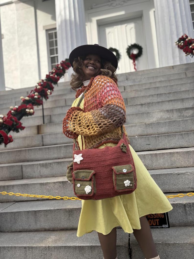 Pu&ograve; includere: Una donna indossa un cardigan di uncinetto multicolore, un vestito giallo e un cappello marrone. Porta una borsa di uncinetto con decorazioni floreali. La borsa ha due tasche grandi e una tasca pi&ugrave; piccola sul davanti.