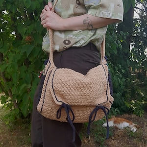 Peut inclure: Un sac &agrave; bandouli&egrave;re en crochet beige avec des bordures et des liens bleu fonc&eacute;. Le sac est port&eacute; sur l'&eacute;paule d'une personne portant une chemise &agrave; motifs et un pantalon marron.