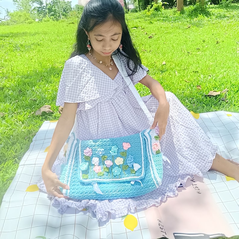 Peut inclure: Un sac en crochet bleu clair avec un motif floral. Le sac a une bandouli&egrave;re blanche et une bordure blanche. Le sac est tenu par une personne portant une robe &agrave; carreaux vichy violet clair.