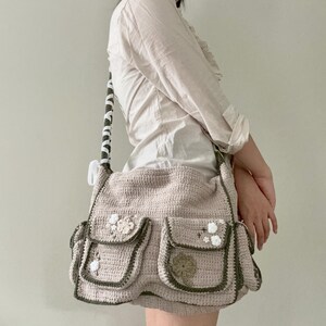 Pu&ograve; includere: Una borsa a tracolla in crochet beige con bordi verdi e decorazioni floreali bianche. La borsa ha pi&ugrave; tasche e una lunga tracolla.