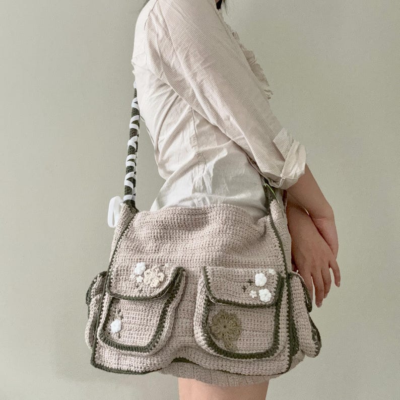 Pu&ograve; includere: Una borsa a tracolla in crochet beige con bordi verdi e decorazioni floreali bianche. La borsa ha pi&ugrave; tasche e una lunga tracolla.