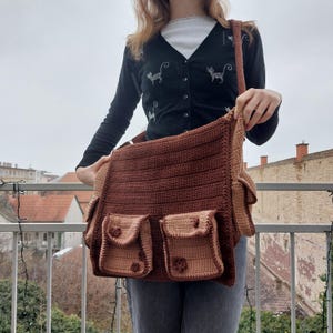 Pu&ograve; includere: Una borsa a tracolla in crochet marrone con pi&ugrave; tasche. La borsa ha una tracolla lunga ed &egrave; realizzata con filato marrone e beige.