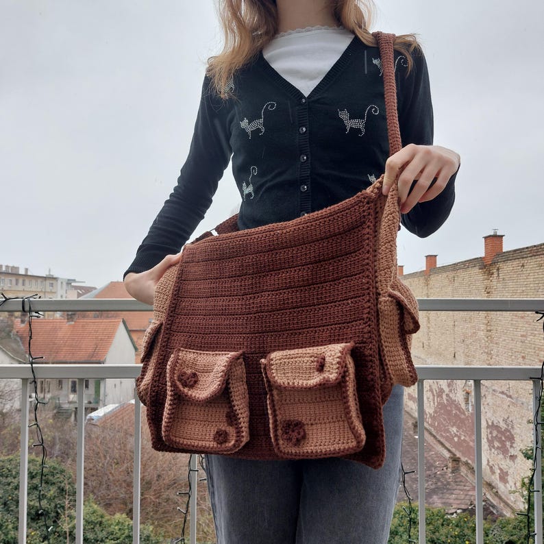 Pu&ograve; includere: Una borsa a tracolla in crochet marrone con pi&ugrave; tasche. La borsa ha una tracolla lunga ed &egrave; realizzata con filato marrone e beige.