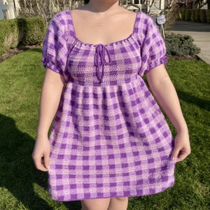 May include: A purple and white gingham crochet dress with a square neckline and short puff sleeves. The dress has a fitted bodice and a flowy skirt.