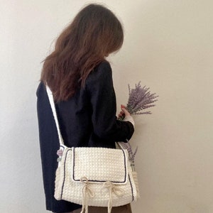 Peut inclure: Un sac &agrave; bandouli&egrave;re en crochet blanc avec une bordure noire et un d&eacute;tail de n&oelig;ud. Le sac a une longue bandouli&egrave;re et est port&eacute; par une personne tenant un bouquet de lavande.