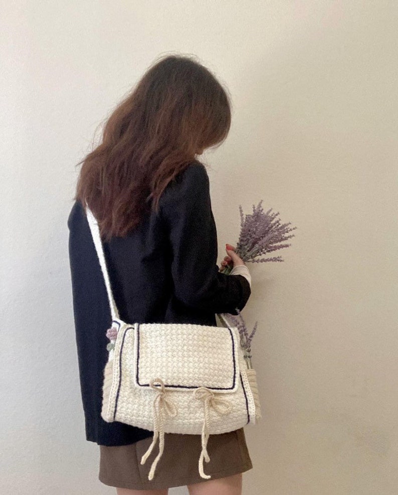 Peut inclure: Un sac &agrave; bandouli&egrave;re en crochet blanc avec une bordure noire et un d&eacute;tail de n&oelig;ud. Le sac a une longue bandouli&egrave;re et est port&eacute; par une personne tenant un bouquet de lavande.