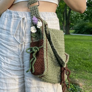 Peut inclure: Un sac &agrave; bandouli&egrave;re en crochet vert avec une poche marron et un embellissement floral. Le sac a une longue bandouli&egrave;re et un pompon marron.