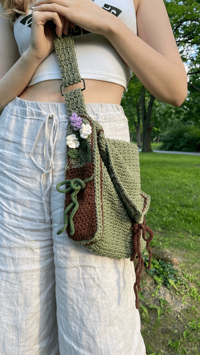 Peut inclure: Un sac &agrave; bandouli&egrave;re en crochet vert avec une poche marron et un embellissement floral. Le sac a une longue bandouli&egrave;re et un pompon marron.