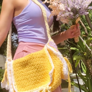 Peut inclure: Un sac &agrave; bandouli&egrave;re en crochet jaune avec des bordures blanches et des glands jaunes. Le sac est port&eacute; par une personne v&ecirc;tue d'un d&eacute;bardeur lavande et d'une jupe rose.