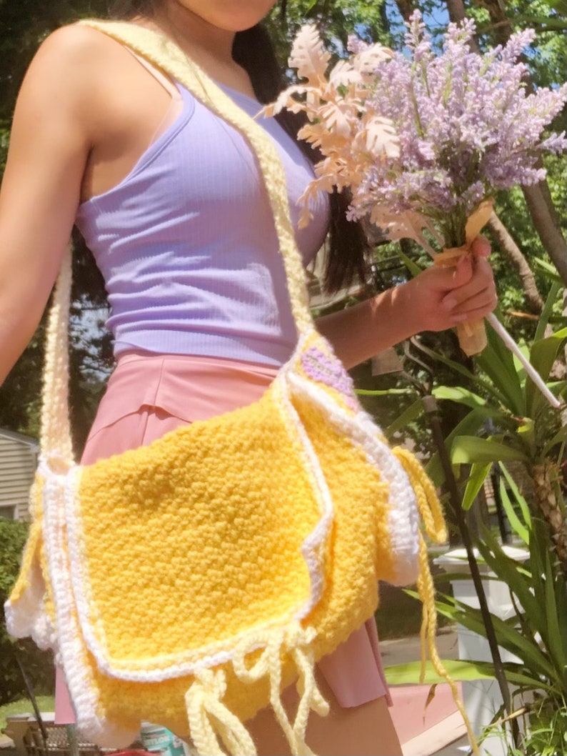 Peut inclure: Un sac &agrave; bandouli&egrave;re en crochet jaune avec des bordures blanches et des glands jaunes. Le sac est port&eacute; par une personne v&ecirc;tue d'un d&eacute;bardeur lavande et d'une jupe rose.