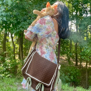 Peut inclure: Une femme v&ecirc;tue d'une robe &agrave; fleurs tient un chat roux dans ses bras. Elle porte un sac &agrave; bandouli&egrave;re en crochet marron avec des finitions vertes et blanches.