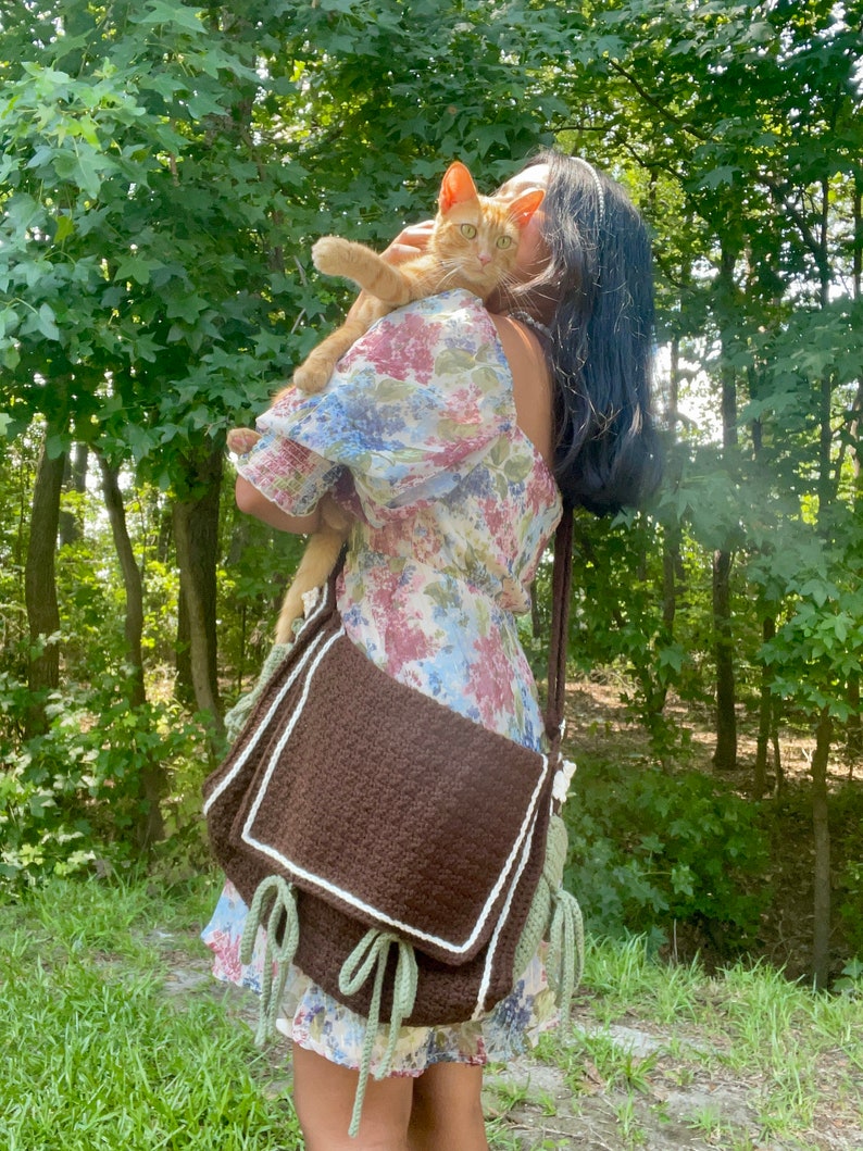 Peut inclure: Une femme v&ecirc;tue d'une robe &agrave; fleurs tient un chat roux dans ses bras. Elle porte un sac &agrave; bandouli&egrave;re en crochet marron avec des finitions vertes et blanches.