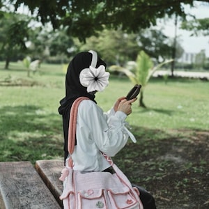 Pu&ograve; includere: Una borsa tote rosa all'uncinetto con pi&ugrave; tasche e una tracolla rosa e bianca all'uncinetto. La borsa &egrave; indossata da una persona seduta su una panchina di legno in un parco.