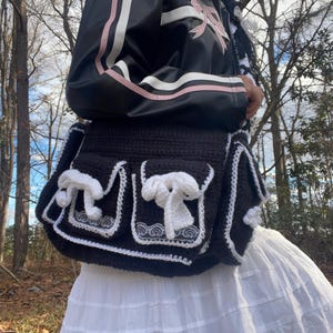 Pu&ograve; includere: Una borsa a uncinetto nera con bordi e fiocchi bianchi. La borsa ha due tasche grandi e una tasca pi&ugrave; piccola nella parte anteriore. La borsa viene indossata da una persona che indossa una giacca nera e rosa e una gonna bianca.