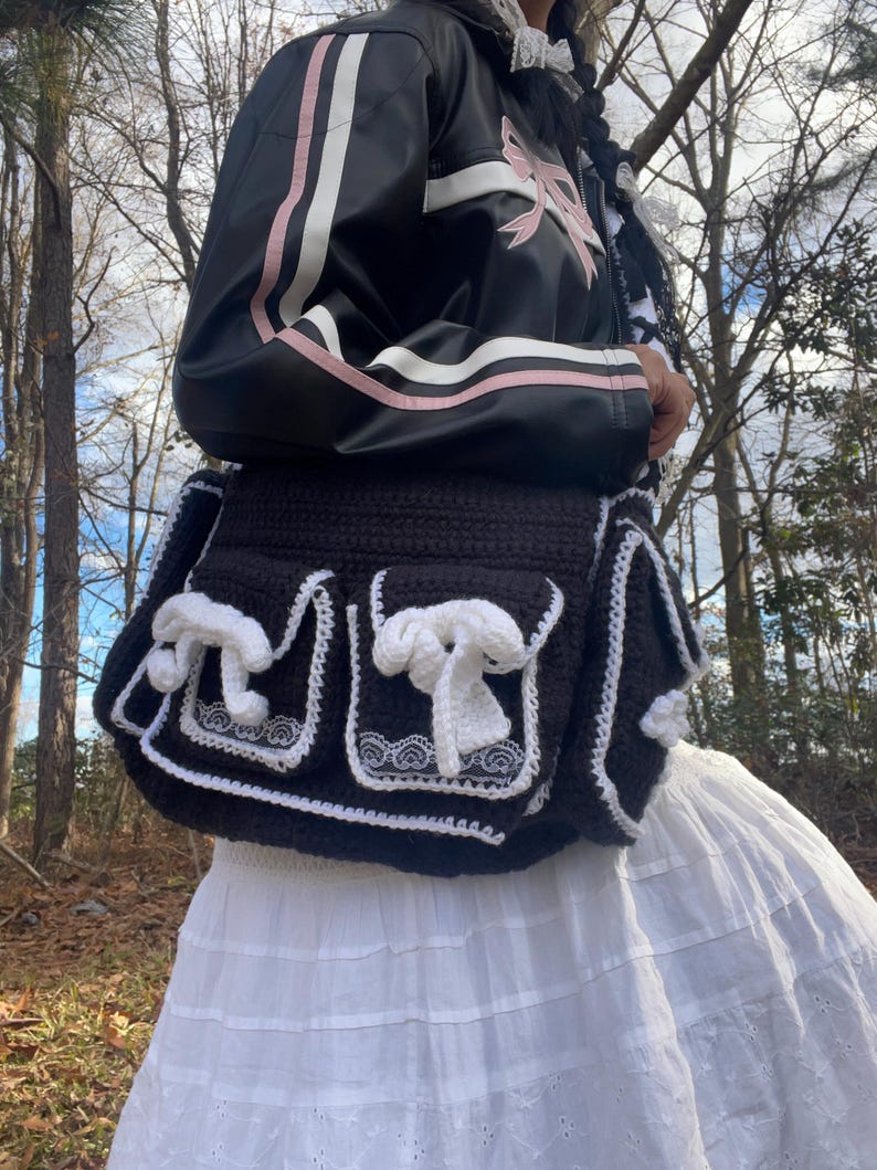 Pu&ograve; includere: Una borsa a uncinetto nera con bordi e fiocchi bianchi. La borsa ha due tasche grandi e una tasca pi&ugrave; piccola nella parte anteriore. La borsa viene indossata da una persona che indossa una giacca nera e rosa e una gonna bianca.