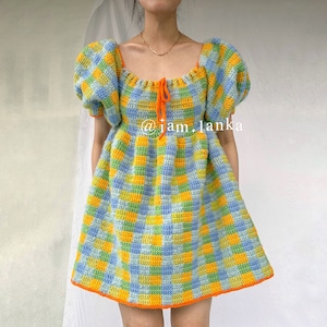 May include: A crochet dress with a checkered pattern in shades of blue, green, and yellow. The dress has a square neckline and short, puffed sleeves. The dress is tied at the neckline with a bright orange ribbon.