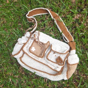 Pu&ograve; includere: Una borsa bianca all'uncinetto con bordi marroni e una tracolla in crochet marrone. La borsa ha pi&ugrave; tasche e un fiore decorativo sul davanti.