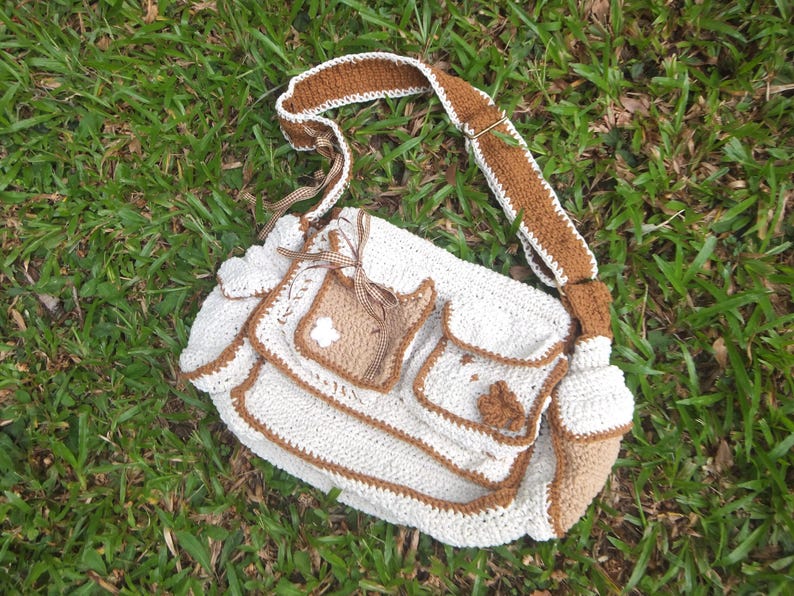 Pu&ograve; includere: Una borsa bianca all'uncinetto con bordi marroni e una tracolla in crochet marrone. La borsa ha pi&ugrave; tasche e un fiore decorativo sul davanti.