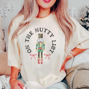 Op de afbeelding: Een crèmekleurig T-shirt met een afbeelding van een notenkraker en de tekst "On the Nutty List".