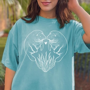 May include: A teal blue t-shirt with a white graphic of two manatees facing each other with a heart between them. The manatees are surrounded by white seaweed.