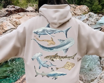 May include: A beige hoodie with a graphic print of various sharks and rays on the back. The hoodie has a drawstring hood and a kangaroo pocket.