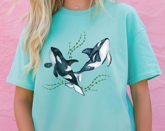 May include: A turquoise blue t-shirt featuring a graphic of two black and white orcas swimming in a sea of green seaweed.