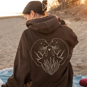May include: A brown hoodie with a graphic of two manatees in a heart shape on the back. The manatees are outlined in white and have a small heart between them. The hoodie is worn by a person sitting on a blanket with a camera and a hat.