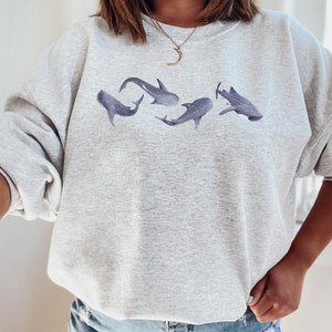 May include: A grey sweatshirt featuring a graphic of five whale sharks in a line. The sweatshirt has a crew neck and long sleeves.