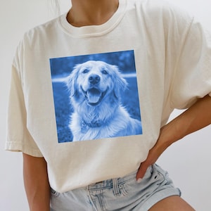 Pode incluir: Camiseta creme com um gráfico quadrado azul de um golden retriever sorrindo. O cão está com a boca aberta, a língua visível e usa uma coleira. A camisa tem mangas curtas e gola redonda.