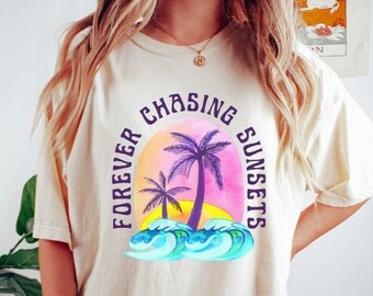 Può includere: Una donna indossa una maglietta beige chiaro con il testo "Forever Chasing Sunsets" e una grafica di palme e onde. La maglietta è oversize e ha una vestibilità rilassata. La donna indossa anche pantaloncini da ciclista verde scuro.