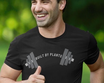 May include: A man wearing a black t-shirt with the text "BUILT BY PLANTS" and a grey barbell graphic. He is running in a grassy field.