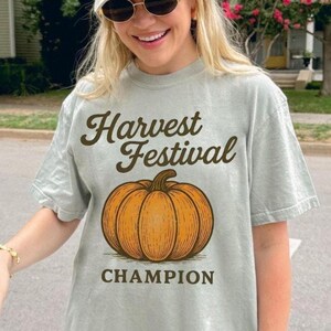 May include: A pale green t-shirt featuring the text "Harvest Festival" above an orange pumpkin illustration and the word "Champion" below. The shirt is worn with a light green cap and sunglasses.