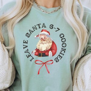 May include: A light green t-shirt with a Santa Claus graphic and the text "LEAVE SANTA 6-7 COOKIES". The Santa illustration features a red suit and hat. A red bow is below the Santa graphic.