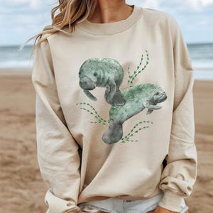 May include: A beige sweatshirt with a watercolor illustration of two manatees swimming in a bed of green seaweed.