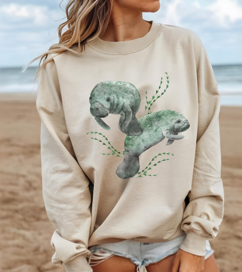 May include: A beige sweatshirt with a watercolor illustration of two manatees swimming in a bed of green seaweed.